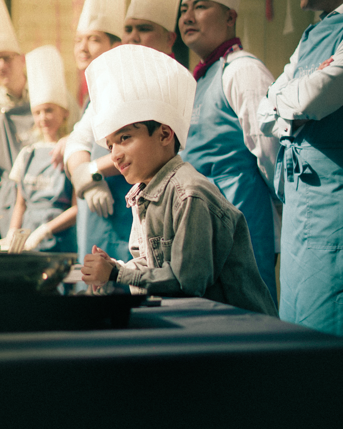 our-story A young boy in a chef hat participates in a lively cooking class with instructors.