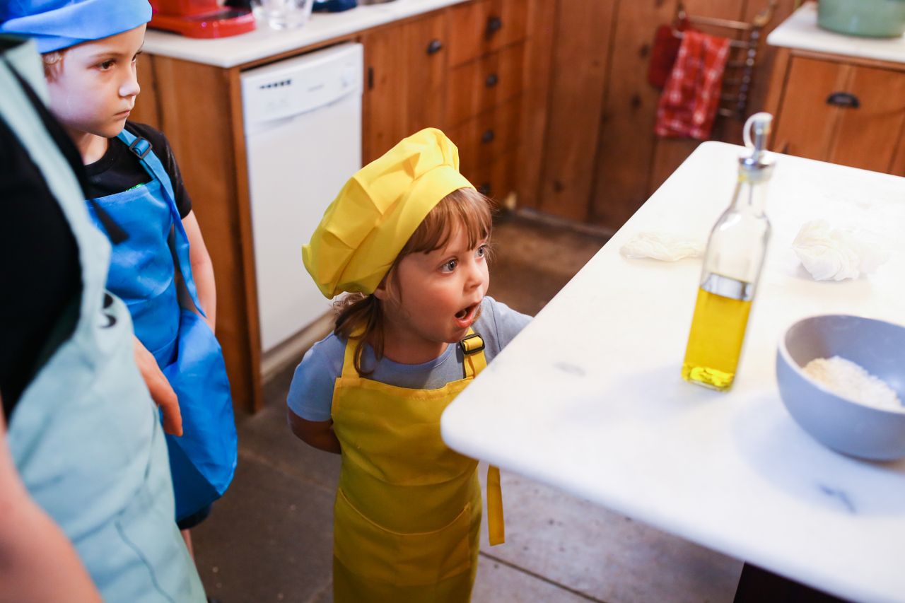 Home Young children in colorful aprons and hats explore cooking at a kitchen counter.